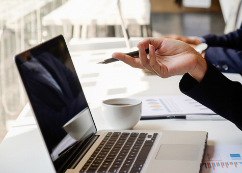 Mano señalando la pantalla de un portátil en una oficina con documentos y una taza de café.